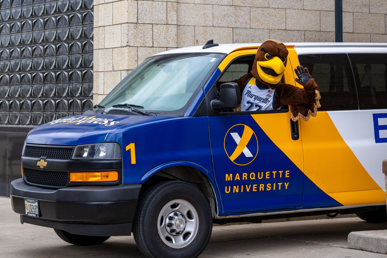 Iggy the Marquette mascot driving an Eagle Express van between the 707 building and Cobeen Hall on the MU campus.